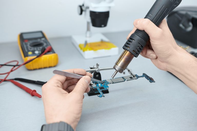 Candid shot of unrecognizable repairman soldering components in motherboard while fixing damaged smart phone, using tweezers and iron. Computer hardware, electronics, repair and upgrade concept